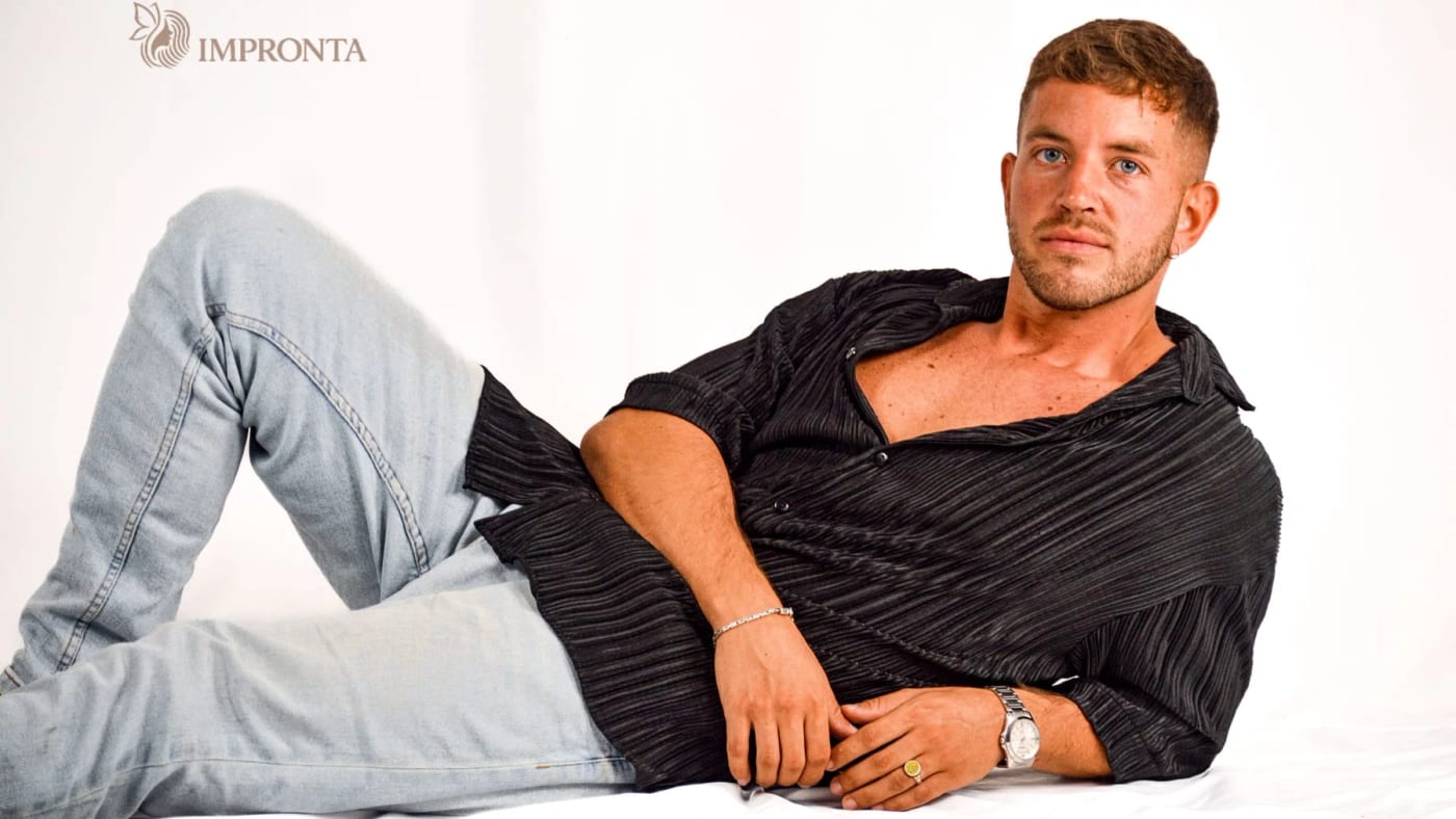 An Impronta model reclining in an open black shirt and light jeans on a white studio backdrop.