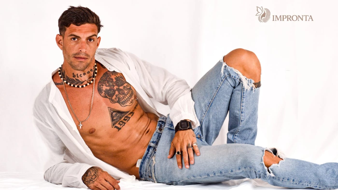 An Impronta model reclining in an open white shirt and light jeans on a white studio backdrop.