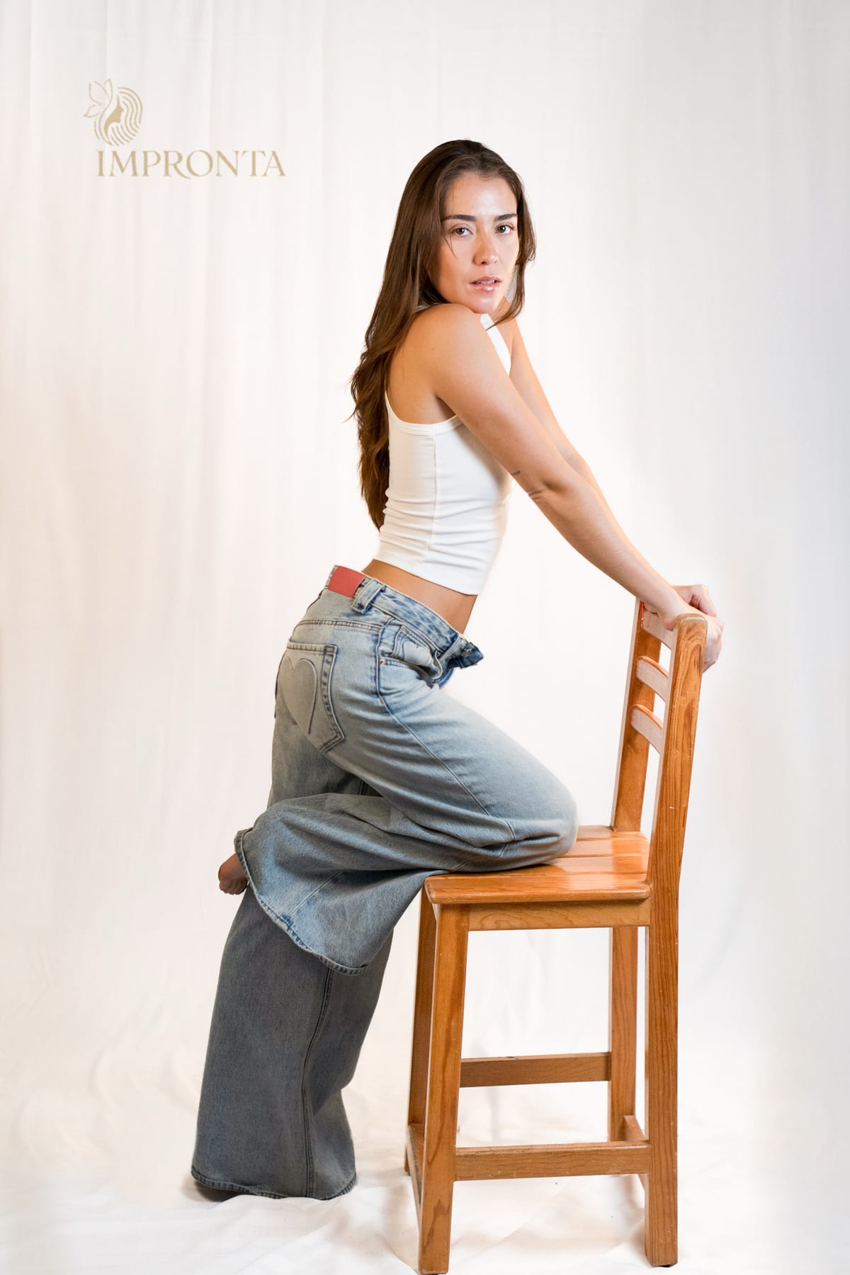 An Impronta model in a white tank top and wide light jeans, leaning against a wooden chair on a white backdrop.