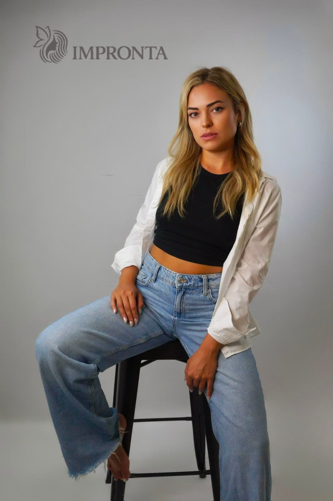 An Impronta model on a stool in a white shirt over a black top and wide jeans, against a grey studio backdrop.