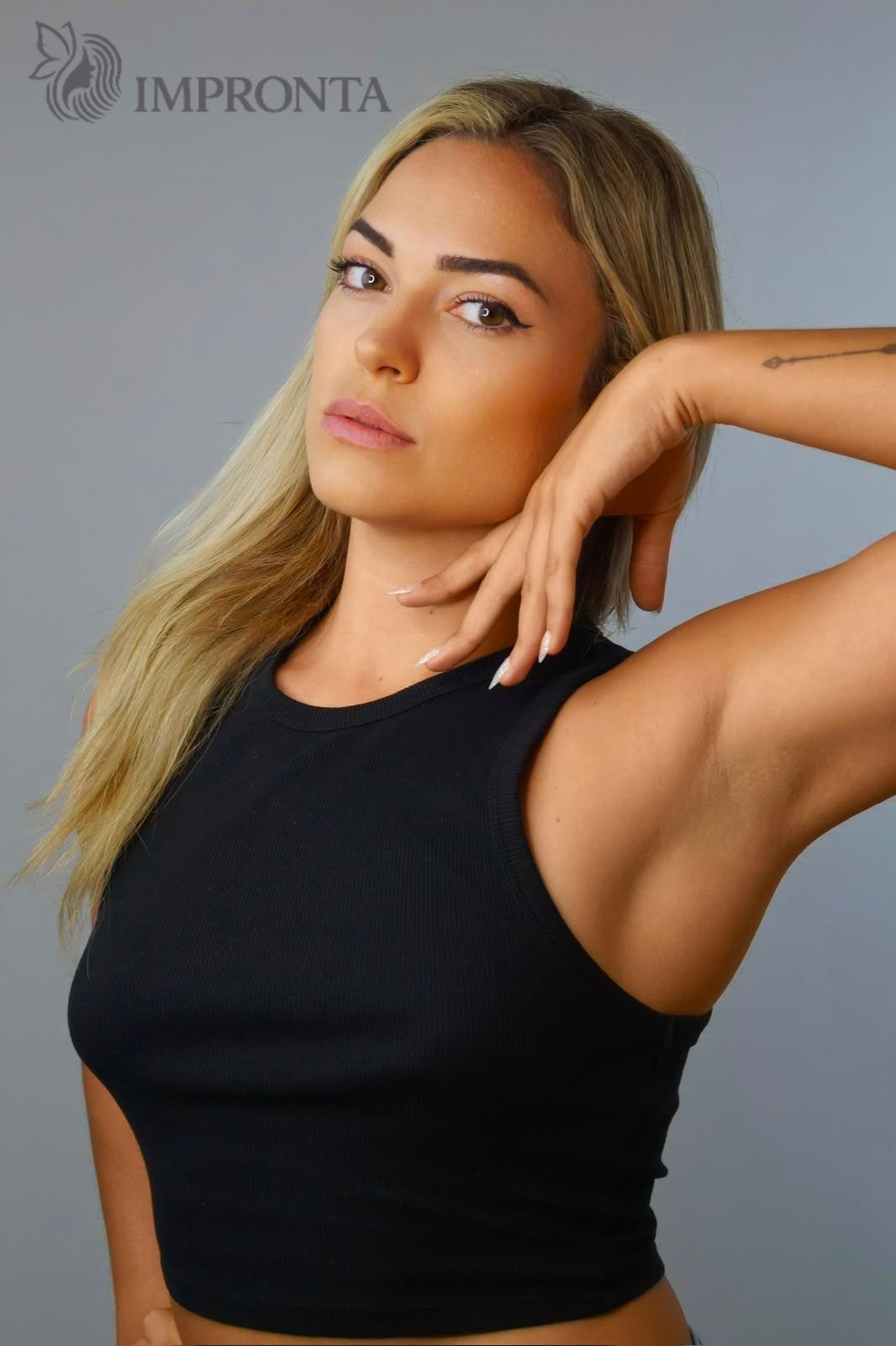 An Impronta model in a black crop top with one hand behind her head, against a grey studio backdrop.