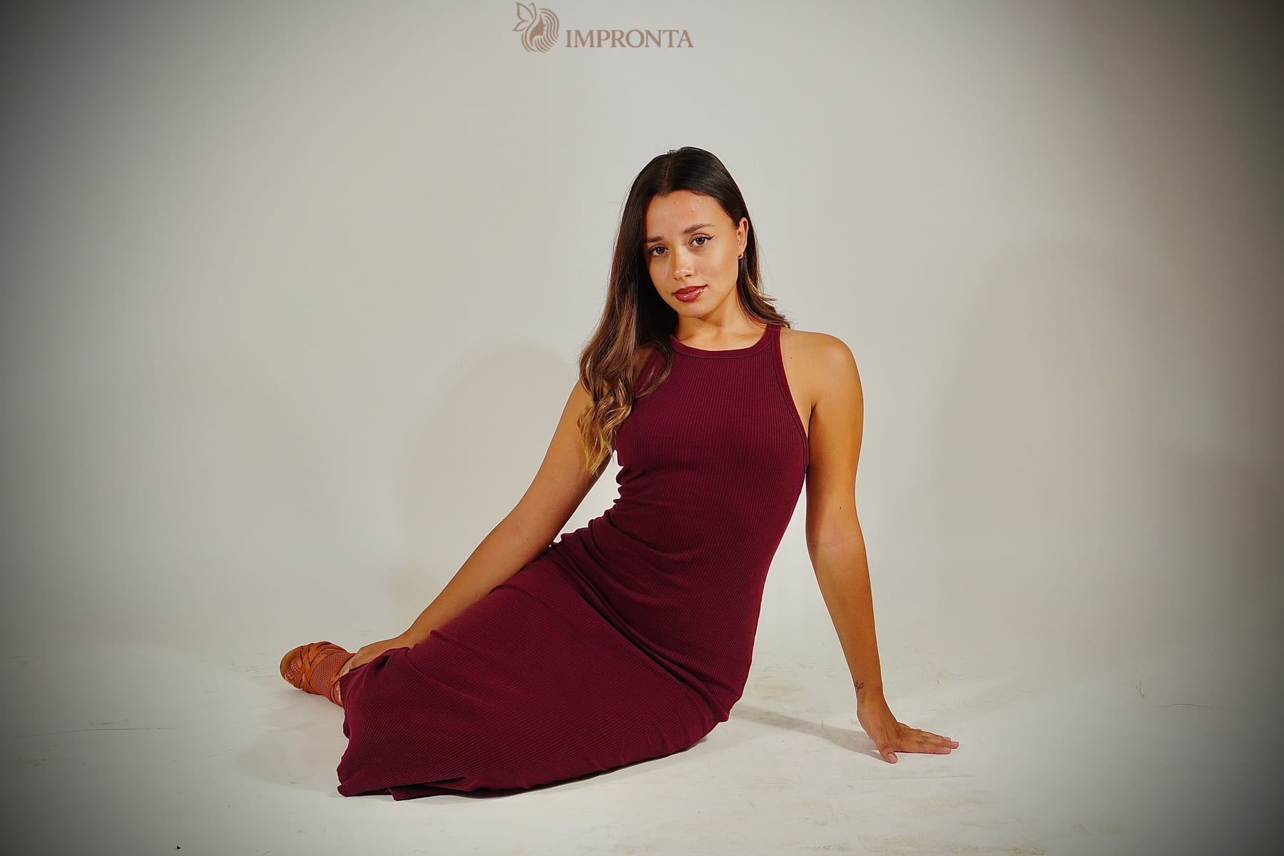 An Impronta model in a burgundy dress, seated on the floor of the agency studio against a white backdrop.