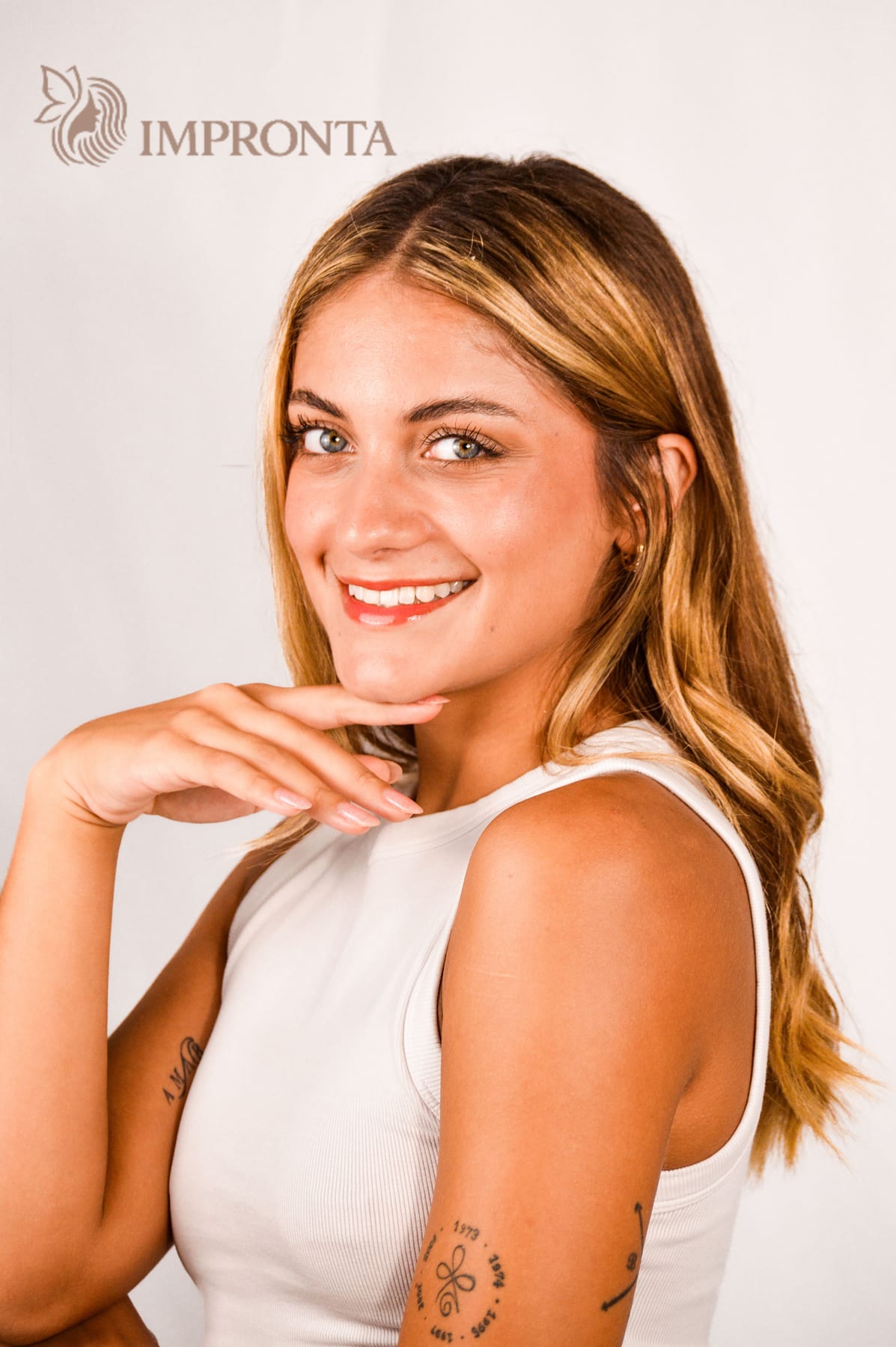 Smiling studio portrait of an Impronta model in a white top, one hand at her chin, against a white backdrop.