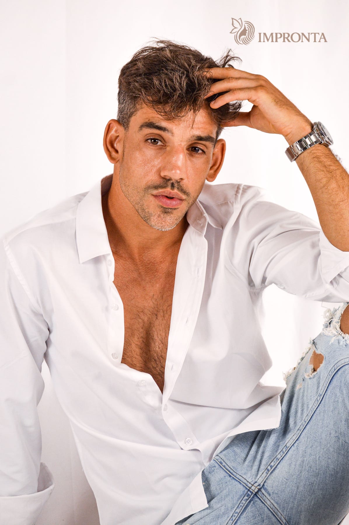 An Impronta model in an open white shirt with one hand in his hair, against a white studio backdrop.