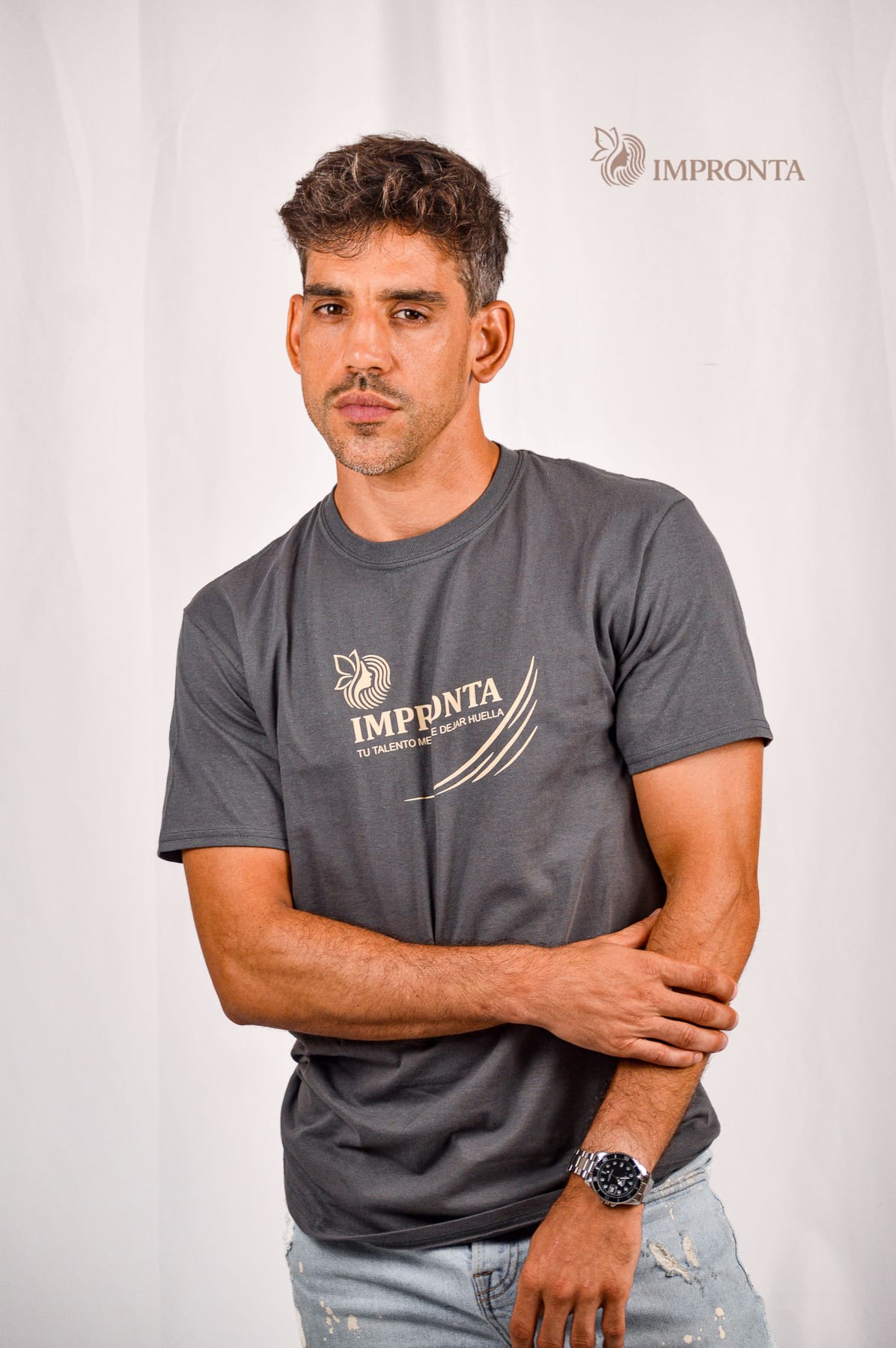 An Impronta model in a grey Impronta t-shirt, arms folded, against a white studio backdrop.
